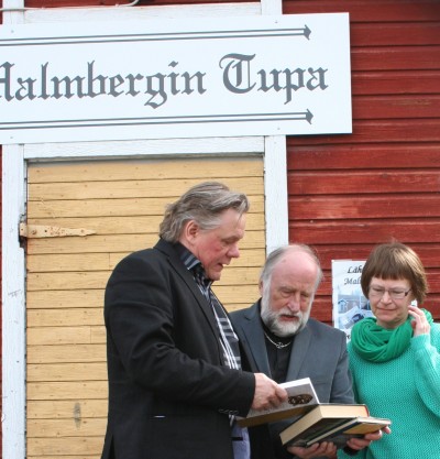 Ohjaaja Raimo Hentunen, kirkkoherra Rauli Junttila ja lhetyssihteeri Anna-Maija Pahkala tutkimassa Malmbergista kertovaa kirjallisuutta Malmbergin tuvan portailla